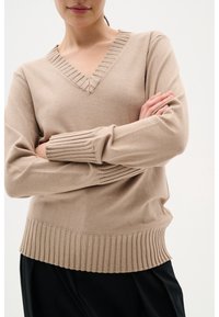 Beige v-ringad tröja med ribbade fållar och ärmslut. Mjukt stickat material med texturerade detaljer längs halsringningen och vertikal ribbning vid ärmsluten.