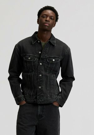 Młody mężczyzna w czarnej kurtce i spodniach z denimu, z rękami w kieszeniach, stojący na tle jednolitego jasnoszarego tła.