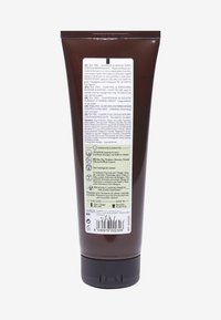 Brun plasttub för tea tree-schampo, med en bred svart kork och olika textetiketter på flera språk på baksidan.