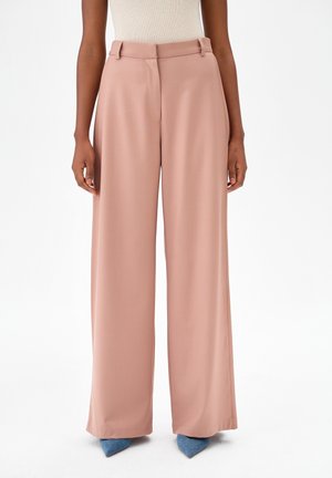 Kobieta ubrana w jasnoróżowe spodnie z wysokim stanem i szerokimi nogawkami, do tego niebieskie buty na szpic oraz biała, bezrękawnikowa, prążkowana bluzka.