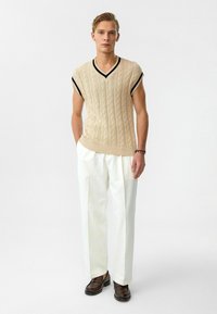 Beige gebreide vest met kabelpatroon, V-hals en zwarte bies, gecombineerd met witte broeken en bruine schoenen. Effen achtergrond.