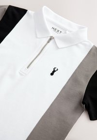 Fehér galléros póló fekete és szürke ujjrészletekkel, cipzáros nyakkal és egy kis fekete homár hímzéssel a mellrészen.