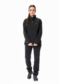 Zwart softshelljack met een hoge kraag en ritssluiting, gecombineerd met zwarte broek en donkere schoenen; model staat rechtop, handen gevouwen.
