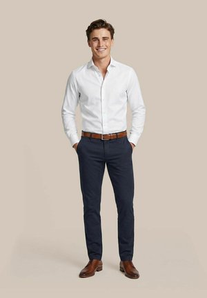 Junger Mann, lächelnd, trägt ein weißes Hemd mit Knöpfen, dunkelblaue Hose, braunen Gürtel und passende braune Anzugschuhe, steht mit den Händen in den Taschen.