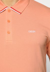 Polo-paita korallioranssista puuvillasta, jossa on jämäkkä rakenne. Siinä on punavalkoinen raidoitettu kaulus ja pieni valkoinen logo rinnassa.