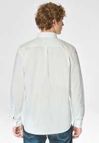 Camicia bianca a maniche lunghe con taglio sartoriale, colletto rigido e polsini abbottonati, caratterizzata da una texture liscia e una leggera lucentezza.