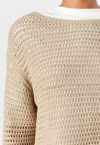 Strickoberteil in offener Maschenstruktur in hellbeige, mit Rundhalsausschnitt und strukturiertem Design, über einem weißen Oberteil mit geradem Saum getragen.