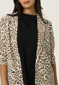 Blazer s uzorkom leopardovih mrlja s krem podlogom, crnim reverom i kratkim rukavima; ima džepove i glatku teksturu. Nositi preko crne majice.