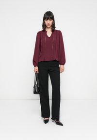 Chemisier bourgogne avec des détails plissés, décolleté en V et manches amples, associé à un pantalon noir ajusté et des escarpins à bout pointu, tenant un sac à main noir.