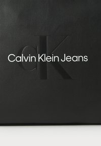 Borsa in pelle nera con logo "CK" in rilievo e testo "Calvin Klein Jeans" in bianco, texture liscia, design minimalista.