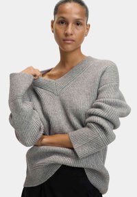 Grauer gerippter Pullover mit V-Ausschnitt und lässiger Passform. Mit voluminösen Ärmeln und einem leicht unebenen Saum. Strukturiertes Strickmuster.