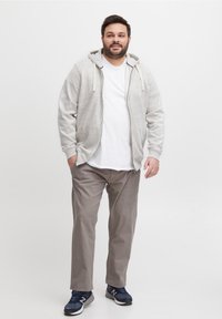 Mann som står iført grå hettejakke, hvit T-skjorte, beige bukser og marineblå sneakers mot en enkel hvit bakgrunn.