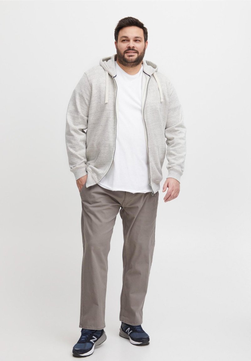 Mann som står iført grå hettejakke, hvit T-skjorte, beige bukser og marineblå sneakers mot en enkel hvit bakgrunn.