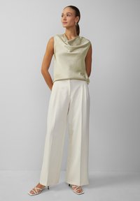 Hellgrünes Cowl-Neck-Top aus glänzendem Stoff, kombiniert mit hochgeschnittenen, weißen weit geschnittenen Hosen. Das Model trägt weiße Riemchen-Heels.