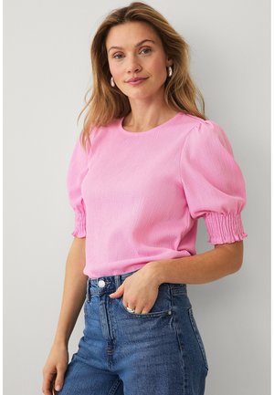 Kvinde med løst, bølget hår, iført sølv øreringe, en lyserød bluse med pufærmer og blå, højtaljede jeans med den ene hånd i lommen.