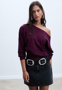 Pull over violet à épaules dénudées avec des manches longues, associé à une jupe en jean noire et une large ceinture noire avec des accents argentés et des œillets.