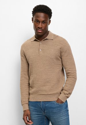 Man draagt een beige gestructureerd poloshirt met lange mouwen en blauwe spijkerbroek, staat met één hand in zijn zak tegen een effen witte achtergrond.