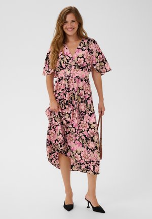 Femme souriante, portant une robe noire à motif floral rose, tenant un sac à main marron, debout en talons noirs sur un fond uni.