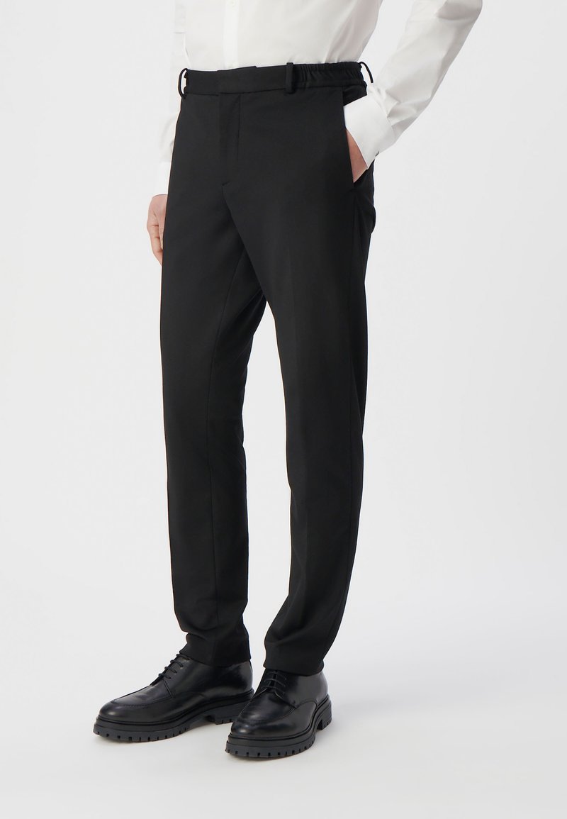 Zwarte broeken met een taps toelopend ontwerp, gemaakt van geweven stof. Voorzien van een elastische tailleband en zijzakken. Gepaard met zwarte leren schoenen.