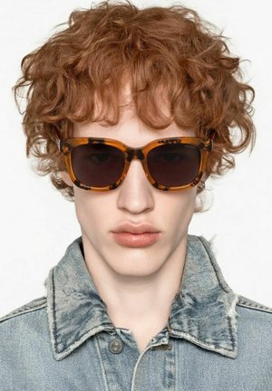 Jeune personne aux cheveux roux bouclés portant des lunettes de soleil écaille de tortue et une veste en denim bleu clair sur un fond uni.