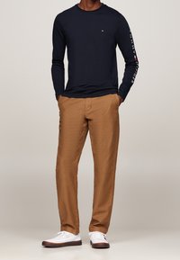 Marineblaue Langarmbluse mit Textlogo auf dem Ärmel, kombiniert mit beigefarbenen Hosen und weißen Sneakers mit braunen Akzenten. Legeres Outfit.