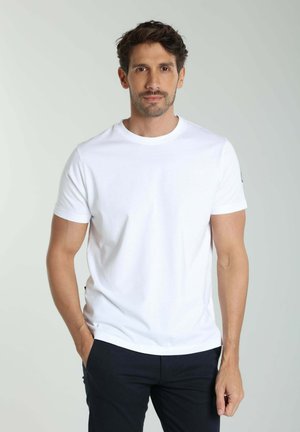 Homme aux cheveux courts et foncés, portant un t-shirt blanc et un pantalon foncé, debout avec une main dans la poche sur un fond clair.