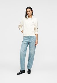 Φωτεινό μπεζ μπουφάν με φερμουάρ και διακριτικό λογότυπο, συνδυασμένο με ανοιχτό μπλε ψηλόμεσο τζιν και μαύρα μποτάκια αστραγάλου. Λείο ύφασμα, άνετη γραμμή.