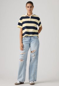 Κίτρινη και ναυτικό μπλε ριγέ πόλο μπλούζα με γιακά· συνδυάζεται με ανοιχτό μπλε σκισμένο τζιν παντελόνι με φαρδιά μπατζάκια και sneakers με καφέ λεπτομέρειες.