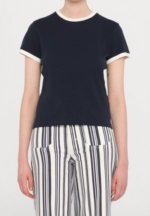 Osoba ubrana w granatową koszulę z krótkim rękawem z białą lamówką oraz białe spodnie z wysokim stanem, ozdobione pionowymi granatowymi paskami i przednimi kieszeniami.