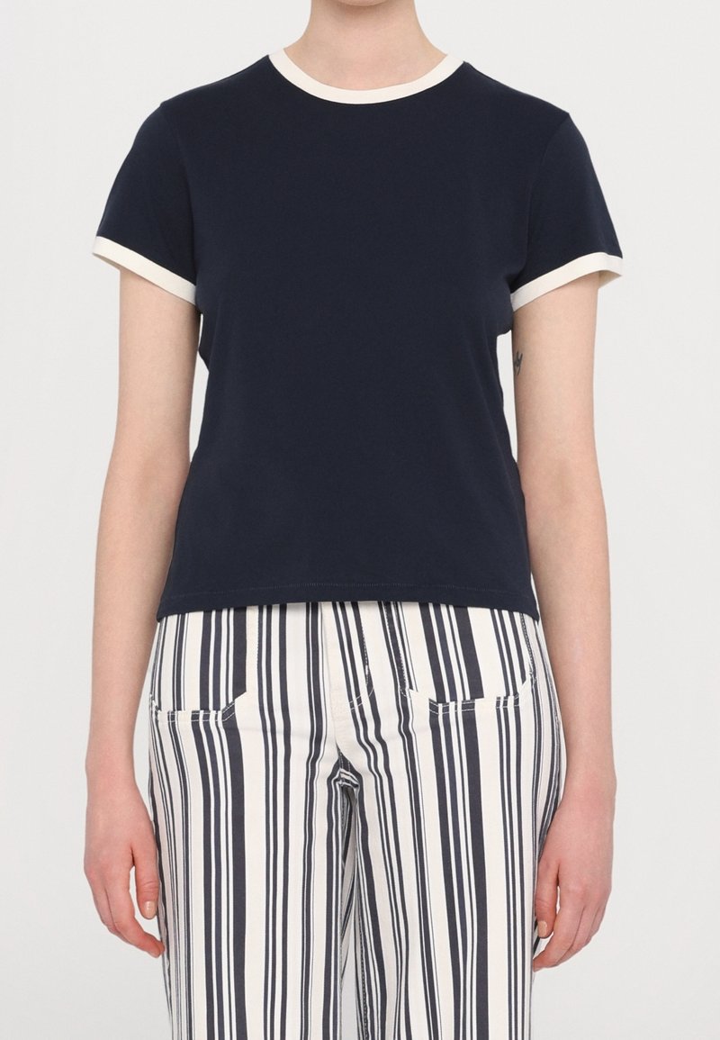 Osoba ubrana w granatową koszulę z krótkim rękawem z białą lamówką oraz białe spodnie z wysokim stanem, ozdobione pionowymi granatowymi paskami i przednimi kieszeniami.