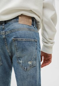Bakåtsikt av en person som bär ljusblå rivna jeans av denim med Pull&Bear läderetikett och vit långärmad skjorta.