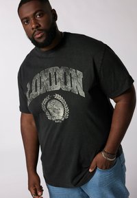 Černé grafické tričko s nápisem "LONDON" v opotřebovaném stříbrném písmu, pod kterým je design vavřínu. Stylizováno s světle modrými džínami.