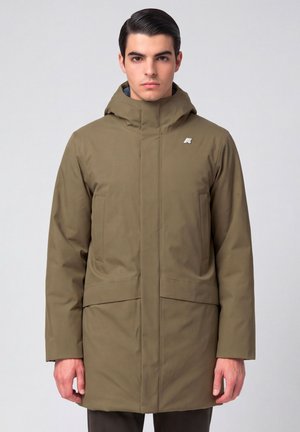 Jonge man die naar voren kijkt, draagt een lange olijfgroene winterjas met capuchon tegen een effen lichte achtergrond.