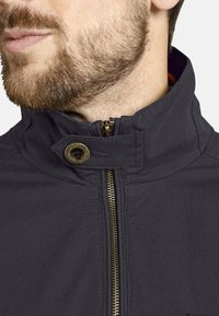 Dark gray zip-up jacket with a high collar, featuring a gold zipper and a buttoned strap closure. Fabric appears smooth with a matte finish.