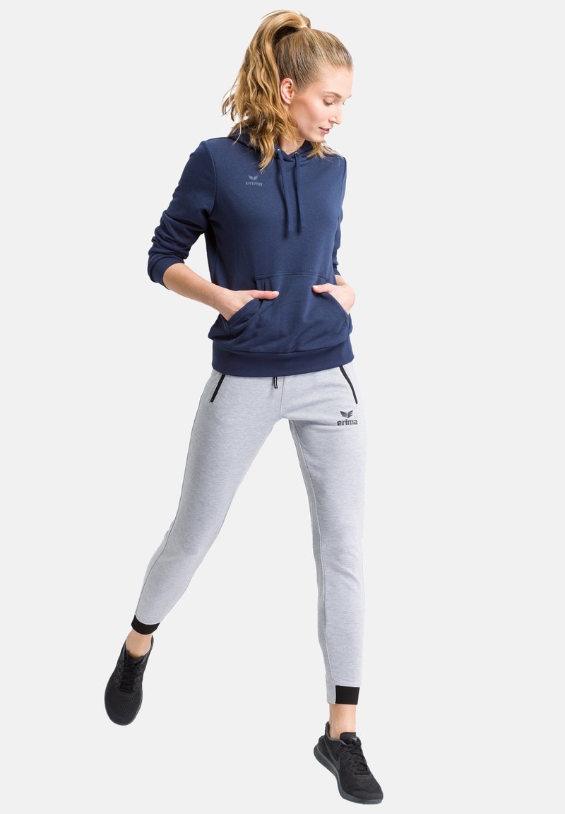 Marineblaues Kapuzensweatshirt mit Kängurutaschen, kombiniert mit hellgrauen, schmal geschnittenen Jogginghosen mit schwarzen Akzenten und Reißverschlusstaschen. Schwarze Sneakers.