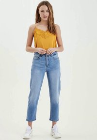 Camiseta amarilla con encaje, combinada con vaqueros de cintura alta de color azul claro. La modelo lleva zapatillas blancas y está de pie frente a un fondo neutro.