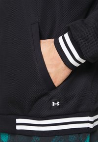 Main dans une poche de veste en maille noire avec un poignet et une ceinture rayés noirs et blancs, arborant une petite étiquette avec le logo Under Armour.