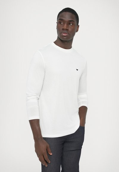 Άσπρη μπλούζα με μακριά μανίκια και στρογγυλή λαιμόκοψη, με μικρό μαύρο λογότυπο στο στήθος. Φορεμένη με σκούρο τζιν παντελόνι. Λείος υφασμάτινος ιστός.