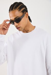 Man with braided hair wearing black sunglasses and white sweatshirt, adjusting sunglasses with his right hand, tattoo visible on neck.