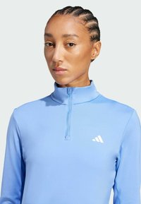 Ljusblå atletisk topp med halvlång dragkedja, tillverkad av ett mjukt material, med en vit logotyp på vänster bröst.