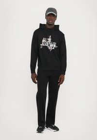 Sweatshirt preto com gráfico "Versace Jeans Couture", combinado com calças pretas e ténis pretos. Apresenta um boné cinza com uma faixa padrão.