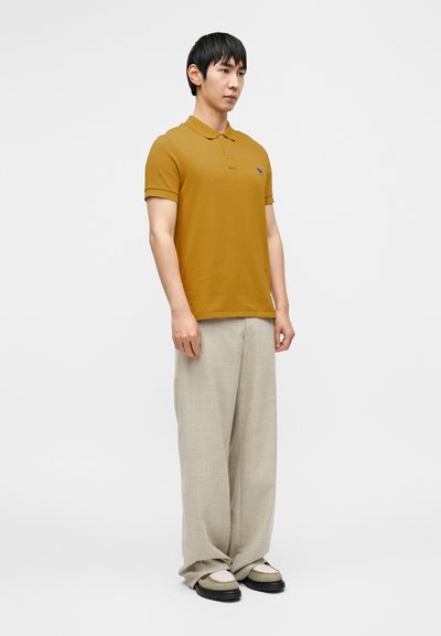 Μουσταρδί κίτρινη πόλο μπλούζα με μικρό λογότυπ ο, συνδυασμένη με ανοιχτό μπεζ παντελόνια με φαρδιά γραμμή και μαύρα αθλητικά παπούτσια με πράσινες λεπτομέρειες.