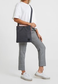 Šedá textilní taška přes rameno s oranžovým zipem a logem, obdélníkový tvar, nošena přes bílé tričko a šedé kostkované kalhoty.