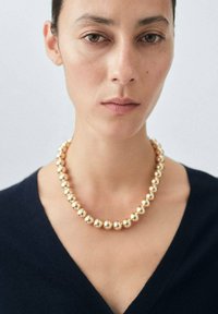 Collana d'oro con perle sferiche lisce. Posizionata attorno al collo, contrasta con un top scuro a V. Design minimalista.