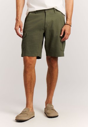 Mężczyzna w oliwkowo-zielonych szortach cargo, beżowych wsuwanych butach oraz białej koszulce, stojący na tle jednolitego jasnego tła.