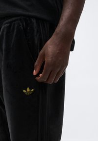 Nahaufnahme einer Hand, die neben einer schwarzen Adidas-Hose mit goldenem Trefoil-Logo am Oberschenkel auf einem schlichten weißen Hintergrund ruht.