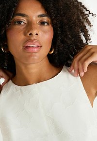 Haut texturé blanc avec motif floral, encolure ronde et léger éclat. Le modèle a des cheveux bouclés et tient l'encolure d'une main.