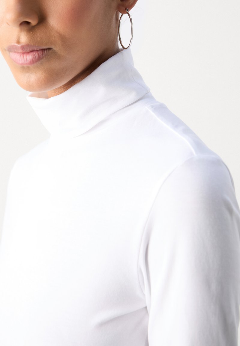 Maglione bianco a collo alto, con vestibilità aderente e superficie liscia, caratterizzato da un colletto alto e maniche sagomate. Orecchino a cerchio in argento visibile.