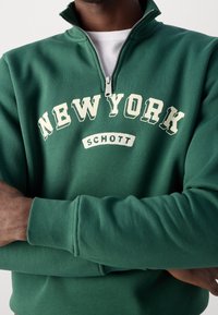 Zelená mikina s vysokým límcem, půlzipem, bílým nápisem "NEW YORK" a logem "SCHOTT". Má žebrované manžety a jemnou texturu.