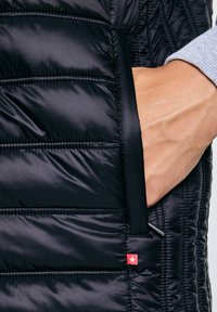 Hand gedeeltelijk in het ritsvak van een zwart doorgestikt jack met horizontale stiksels en een klein rood label met een witte ster.